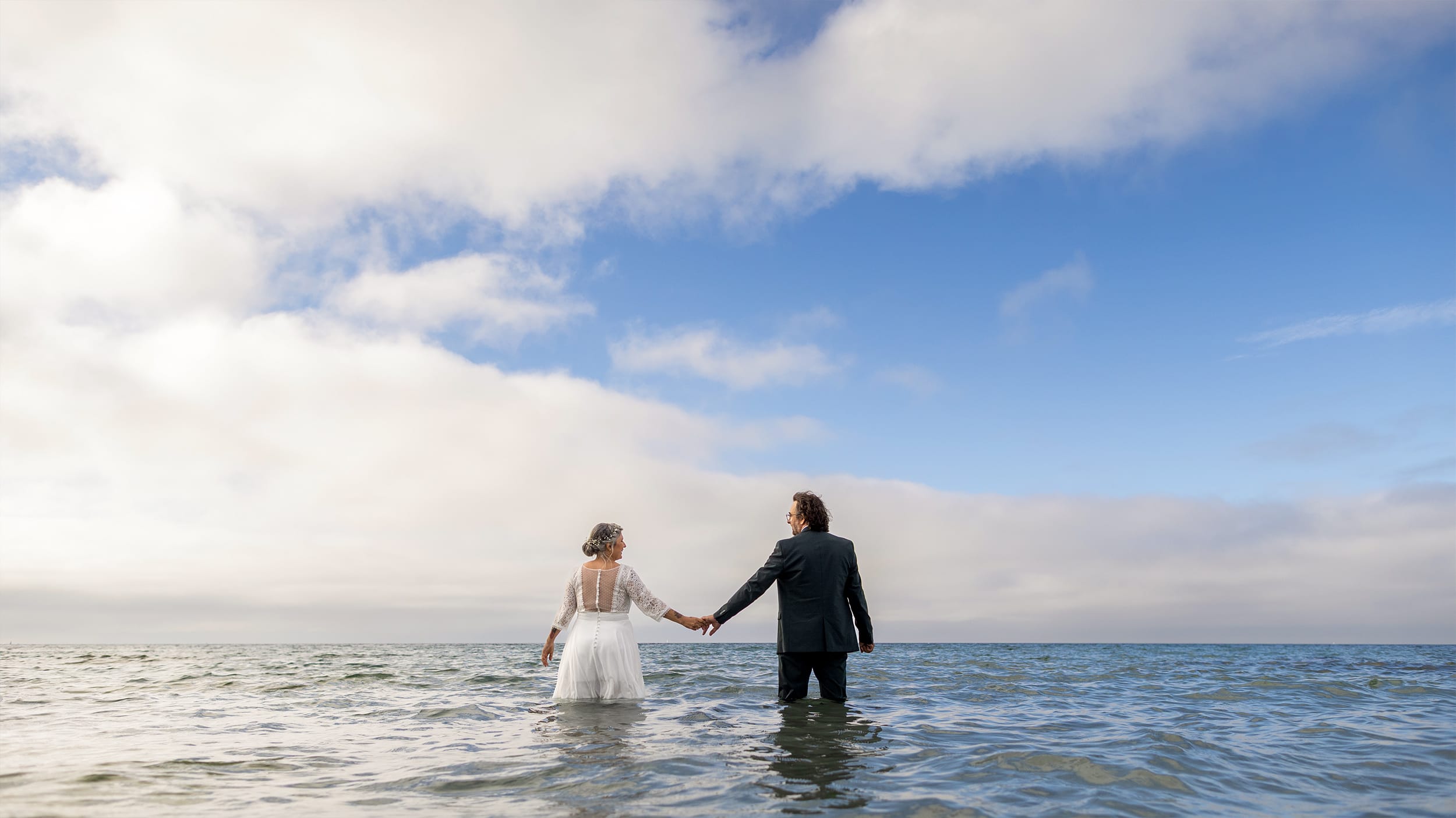 un couple en tenue de mariage se tienne la main dans l'ocean breton face a un ciel de fin de journée.